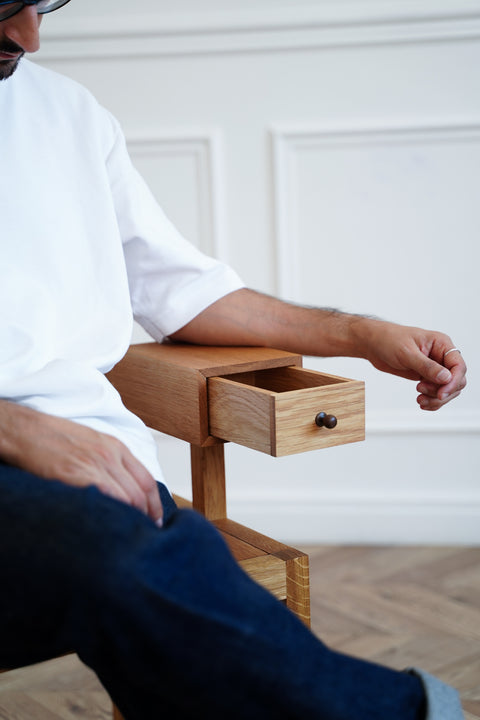 Person sitting on a wooden chair with a drawer, arm rested on the chair's arm, against a white paneled wall.